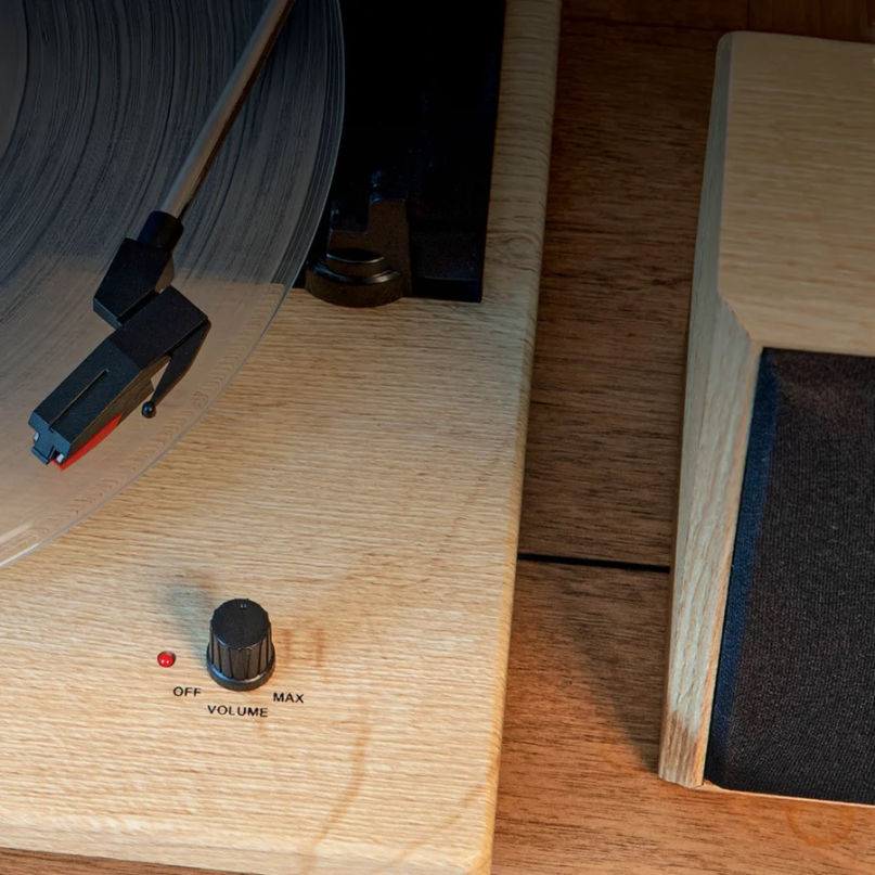Crosley Brio Record Player Shelf System With Speakers A close-up of the crosley brio record player shelf system with speakers playing a vinyl record, showing the tonearm, volume knob, and part of the matching wooden speaker on a wooden surface.